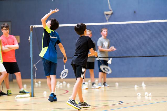 Gruppe von Kindern beim Badmintontraining in einer Sporthalle, mit Schlägern und Federbällen auf dem Boden.