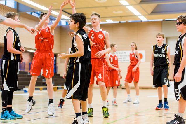 Zwei Basketballteams in action, ein Spieler springt zum Korb, während andere sich um ihn gruppieren.