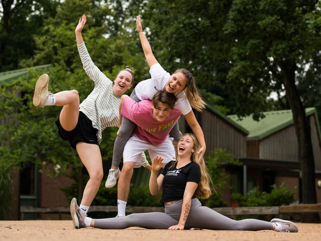 Vier junge Frauen posieren fröhlich auf einer sandigen Fläche, eine sitzt im Spagat, während die anderen sie umarmen.