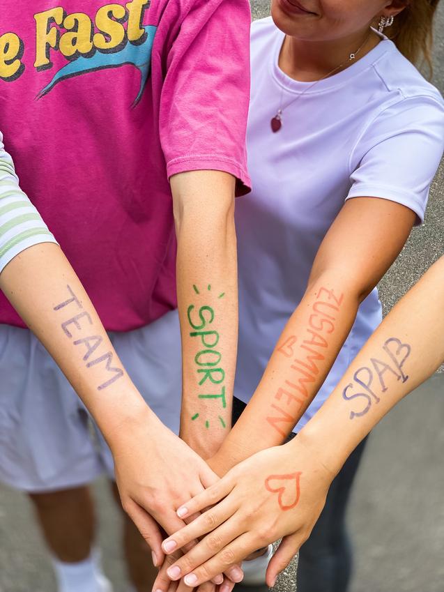 Vier Hände mit bunten Schriftzügen "TEAM", "SPORT", "ZUSAMMEN" und "SPASS" liegen übereinander auf einem grauen Untergrund.