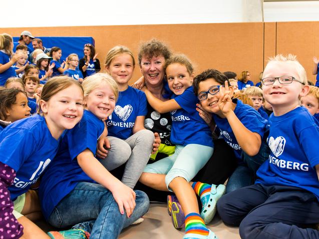 Gruppenkinder in blauen T-Shirts umarmen eine Frau, die lächelt, in einer Sporthalle voller Menschen.