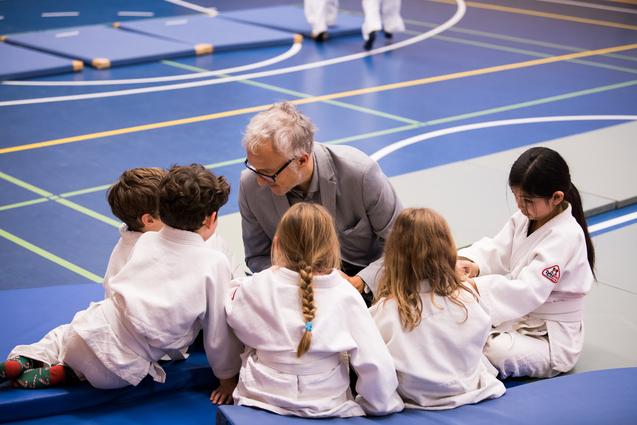 Ein Trainer spricht mit einer Gruppe von fünf Kindern in Judoanzügen auf einer Sportmatte.