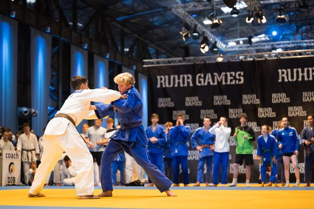 Zwei Jugendliche im Judoanzug kämpfen auf einer gelben Matte bei den Ruhr Games, während Zuschauer im Hintergrund beobachten.