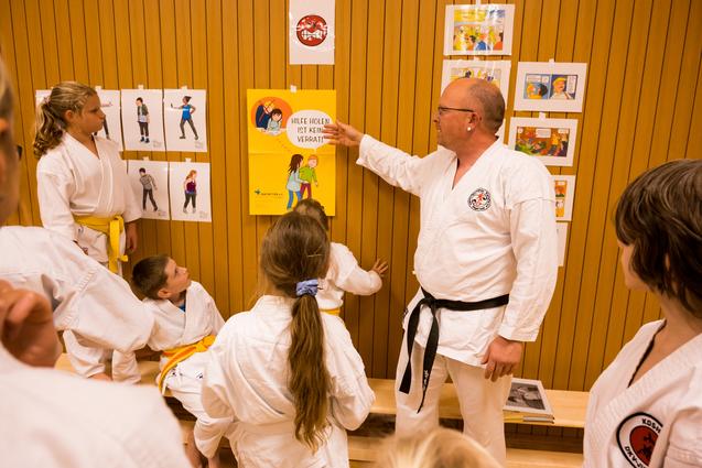 Ein Trainer zeigt Kindern an einer Wand mit Illustrationen eine Sicherheitsbotschaft. Einige Kinder hören aufmerksam zu.