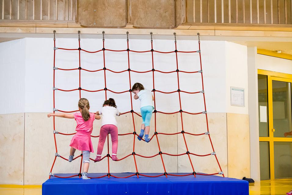 Drei Kinder klettern an einem roten Kletternetz in einer Turnhalle, mit blauer Matte darunter.