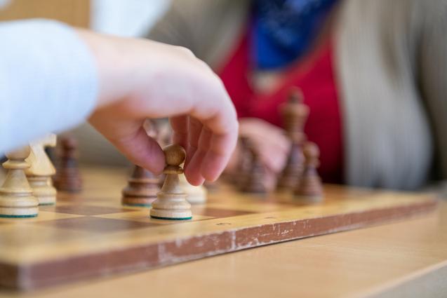 Nahaufnahme einer Hand, die einen Schachbauern auf einem Holzschachbrett hebt, im Hintergrund weitere Figuren sichtbar.