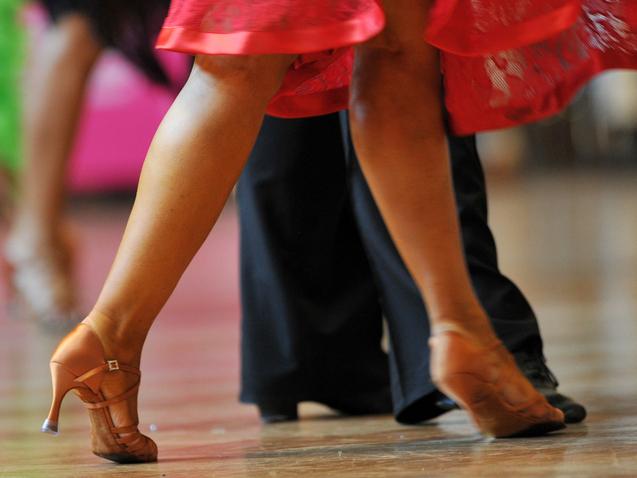 Tanzende Beine in eleganten Schuhen, während sich ein rotes Kleid dreht, vor unscharfem Hintergrund.