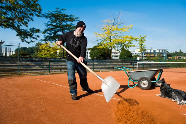 Mann mit Schaufel bearbeitet einen roten Sandsportplatz, während ein Hund daneben liegt und eine Schubkarre steht.